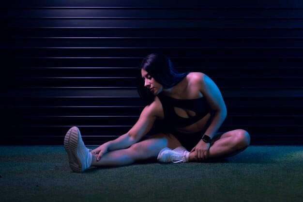A person performing a half-kneeling ankle mobility stretch, gently leaning forward, with a focus on their ankle and lower leg, in a brightly lit gym.