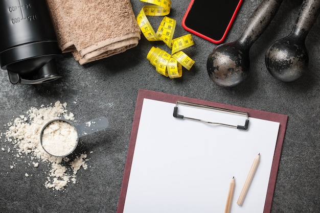 A detailed flat lay of a training journal, a pen, and a protein shaker on a gym mat, representing meticulous planning and post-workout recovery.